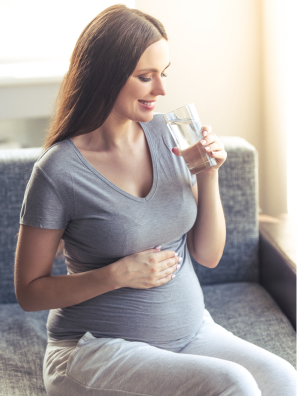 Beautiful pregnant woman is drinking water, keeping one hand on her tummy and smiling while sitting on the couch at home