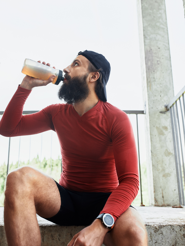 Side view of active bearded sportsman drinking water from glass bottle while having rest and sitting on stairs in bright day